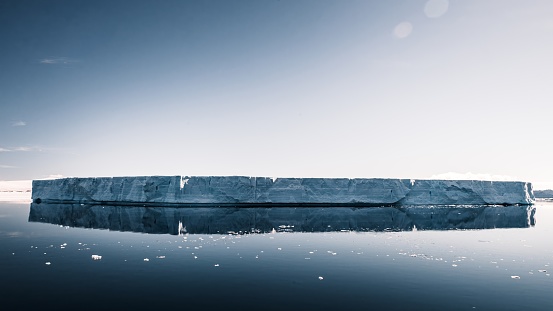 Large Tabular Iceberg floating on the Antarctic Ocean at the coast of Antarctica Peninsula in the sun under sunny cloudless sky. Tabular Iceberg mirroring in the tranquil, calm Antarctica Ocean. Slightly blue toned. Antarctic Ocean, Coast of Antarctica.