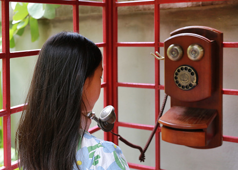 Asian young girl child using dial telephone booth. Kid call on the retro vintage red phone booth, Rear view.