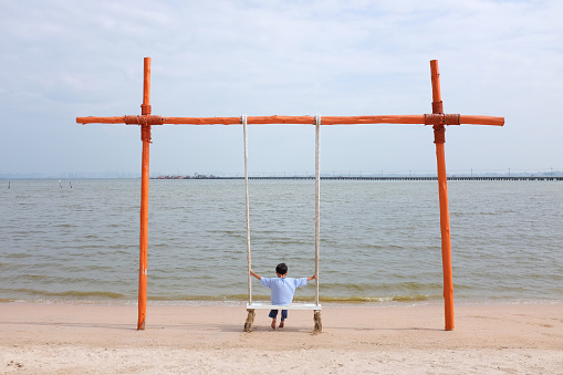 Peaceful Asian boy child sitting on swing at the beach. Kid relax on summer holiday vacation.