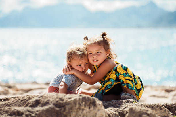 An adorable three year old Eurasian girl affectionately hugs her one year old brother as they enjoy playing outside together in Hawaii, with the ocean visible in the background.