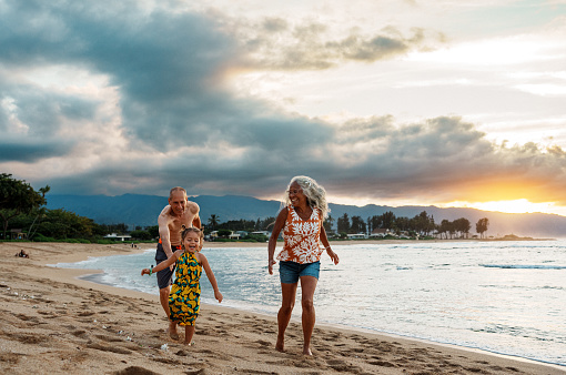 An adorable three year old Eurasian girl of Hawaiian and Chinese descent runs along a sandy beach in Hawaii at sunset and laughs while with her active grandparents playfully chase her.