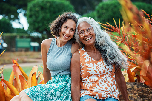 Portrait of a beautiful and healthy senior woman of Hawaiian and Chinese descent and her Eurasian adult daughter smiling directly at the camera while sitting together outside surrounded by orange and yellow tropical foliage in Hawaii.