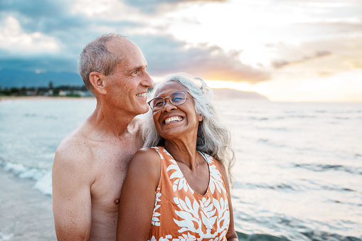 A beautiful multiracial senior woman of Hawaiian and Chinese descent and her Caucasian husband affectionately embrace and smile while at the beach in Hawaii at sunset