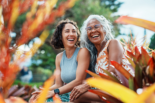 A beautiful and healthy senior woman of Hawaiian and Chinese descent laughs while enjoying quality time outdoors with her Eurasian adult daughter at a park in Hawaii with beautiful tropical landscaping.