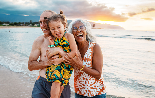 An cute Eurasian three year old girl of Hawaiian and Chinese descent laughs as her grandmother and grandfather affectionately and playfully hold her and pose for a photo while playing at the beach in Hawaii at sunset.