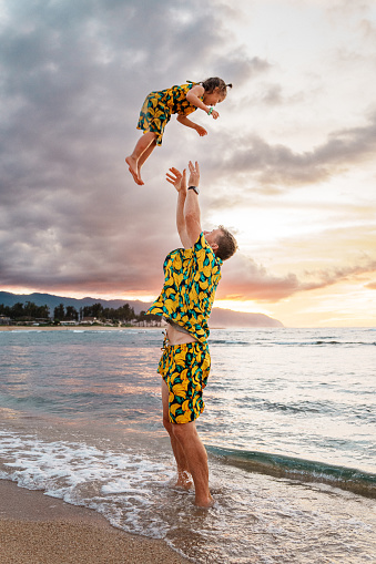 An active young dad playfully tosses his daughter up into the air while playing at the beach in Hawaii at sunset. The father and daughter are wearing matching outfits featuring a banana print.
