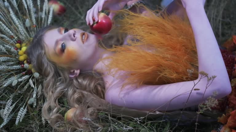 Young woman in autumn designer outfit of flowers, leaves and spikelets lying in the grass among scattered apples