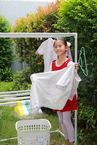 Asian girl child helping to do dry the clothes at the garden near house. Kid hanging cloth on a clothesline.