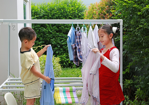 Portrait of Asian girl and boy helping to do dry the clothes at garden near house. Sister and little brother are help to housework. Kids hanging the laundry on a clothesline rail.