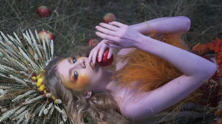 Young woman in autumn designer outfit of flowers, leaves and spikelets lying in the grass among scattered apples