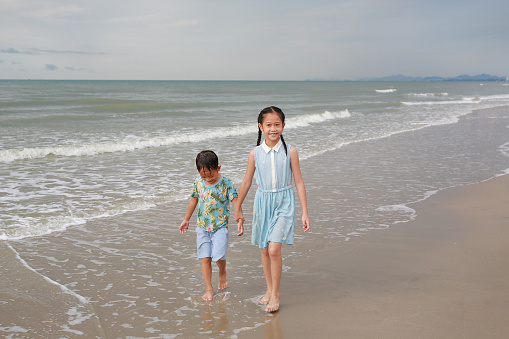 Asian young sister and little brother walking together on tropical sand beach at sunrise. Happy family boy and girl enjoy in summer holiday.