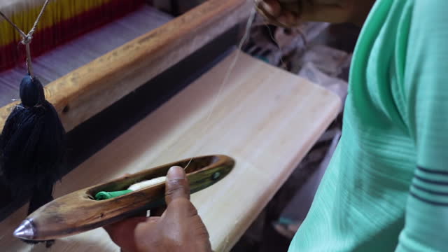 A young man worker works on a machine using yarn on a wooden boat shuttle to make silk cloth