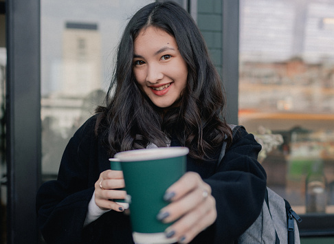 Portrait of cute Korean girl with two coffee in paper cups in cafe near window. Teenage girl having cappuccino and looks at the camera. Life in a big city