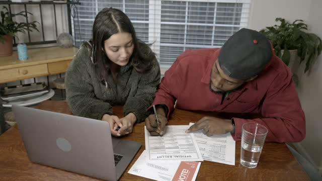 Young Mixed Race Couple Hispanic and African American Sitting at a Kitchen Table Researching Issues and Candidates and Filling Out a Mail In Voting Ballot