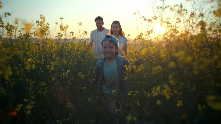 Playing, parents and kid in field of flowers in countryside with sunset sky, freedom or bonding. Family adventure on farm with happy mom, dad and boy walk in nature together with smile, love and fun.