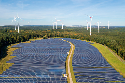 Solar power station and wind power plant surrounded by forest under blue sky, aerial view.