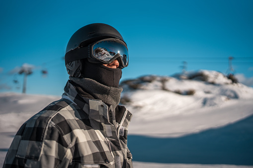 A mid adult Asian male dressed in winter ski attire, including helmet and goggles, standing in a snowy mountain resort with ski lifts in the background.