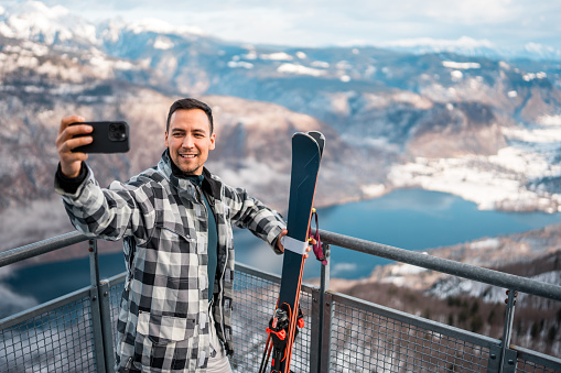 A mid adult Asian male skier captures a selfie with a smartphone while smiling, wearing winter sports gear, with snow-covered mountains and a lake in the background.