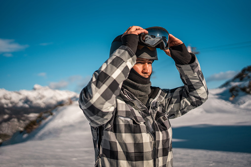 A mid adult Asian male is preparing for skiing by securing his helmet, dressed in a plaid jacket and scarf, with snow-covered mountains in the background.