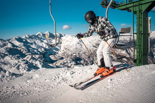 An Asian male in mid-adulthood getting off a ski lift on a a snowy mountain slope, wearing professional ski gear and a helmet near a ski lift, with a clear blue sky and rugged peaks in the background.