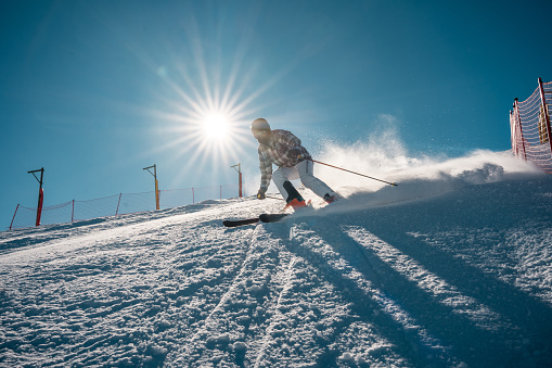 A mid adult Asian male skier is actively descending a snowy mountain slope. He is wearing winter sports attire with ski equipment, demonstrating motion and skill against a bright, sunlit sky.