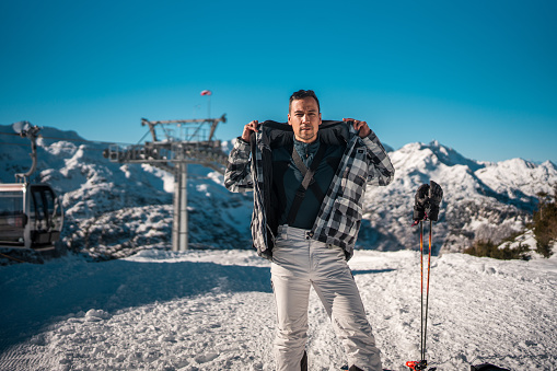 A mid adult Asian male stands in snow with ski equipment, wearing casual ski wear and sunglasses, with a ski lift and mountains in the background.