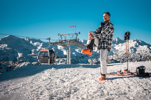 A mid adult Asian male stands on a snow-covered slope, getting ready to ski. He is holding a ski boot, with skis and a helmet placed on the ground next to him, against a backdrop of a clear blue sky and mountain scenery.