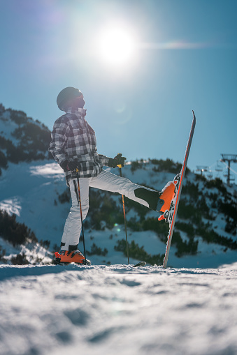 A mid-adult Asian male takes a break from skiing, standing with equipment on a snowy mountain slope under a clear blue sky, wearing winter sportswear and protective gear.
