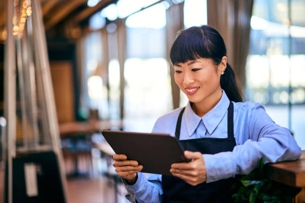 A smiling black-haired Asian waitress, holding a digital menu, dressed in a uniform.