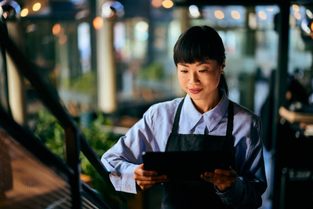 istock An Asian waitress, dressed in a uniform, standing on the stairs, using a digital menu. 1992043499
