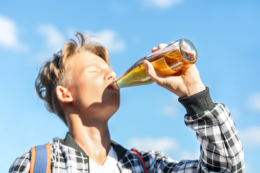 Joyful boy drinking lemonade from a glass bottle against a beautiful sky backdrop. He wears a plaid shirt and a backpack. Vibrant colors
