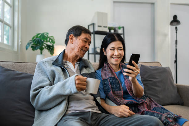 istock Retired elderly couple sits on couch drink tea and using mobile together and relax in their home. Senior Activity Concept 1992079114