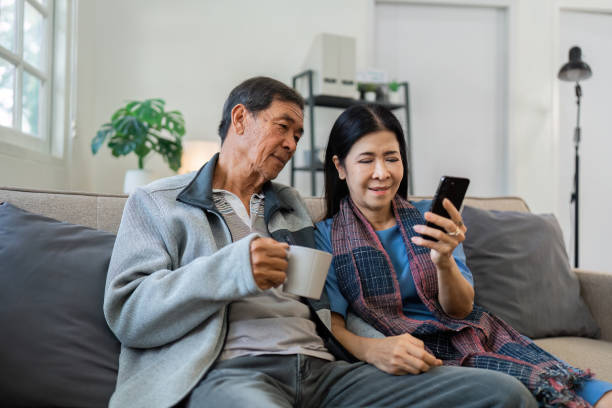 istock Retired elderly couple sits on couch drink tea and using mobile together and relax in their home. Senior Activity Concept 1992092916