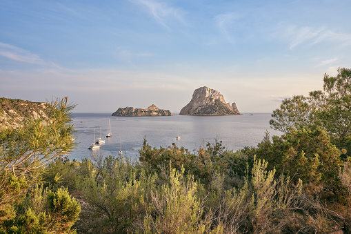 Wide-angle view of the bay of Cala d'Hort, located on the southern coast of Ibiza, renowned for its crystal-clear waters and its scenic view on the iconic rocky isles of Es Vedrà and Es Vedranell. The delicate warm light of a Mediterranean summer sunset, picturesque clouds, sailing boats rocking gently on the water, colourful cliffs covered with lush pine trees and a wide variety of Mediterranean shrubs and plants — juniper, rosemary and thyme among many others. High level of detail, natural rendition, realistic feel. Developed from RAW.