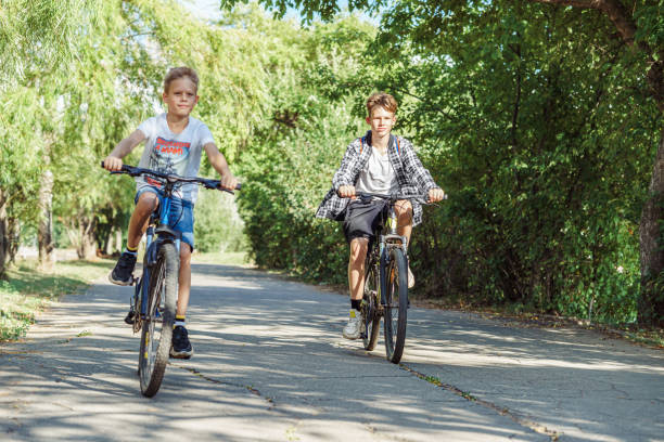 two joyful boys riding bicycles on a bike path in park with lush foliage on a warm summer day - ten speed bicycle imagens e fotografias de stock