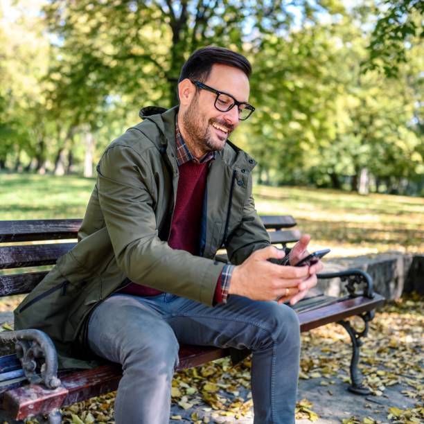 istock Smiling man sitting at park bench and using smartphone 1992113891