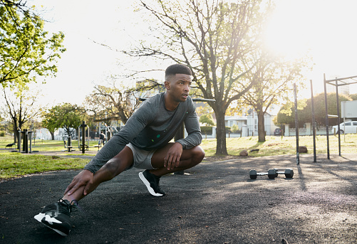 Young black man wearing sports clothing doing stretches in squatting position next to dumbbells at park