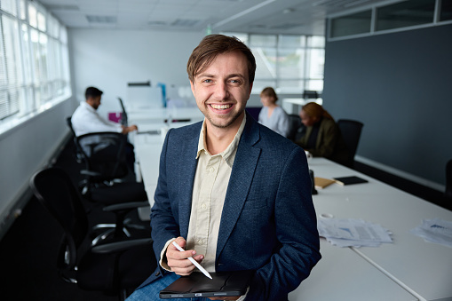 Happy young businessman wearing businesswear sitting on desk with digital tablet and digitized pen in office