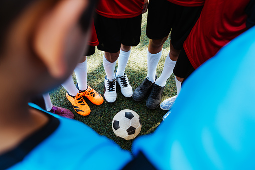 A close-up of young soccer players' feet in a circle around a soccer ball on the turf.