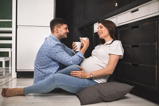 Young pregnant couple in the kitchen, cuddling, preparing food and rejoicing in anticipation of the baby. Concept new life, young family
