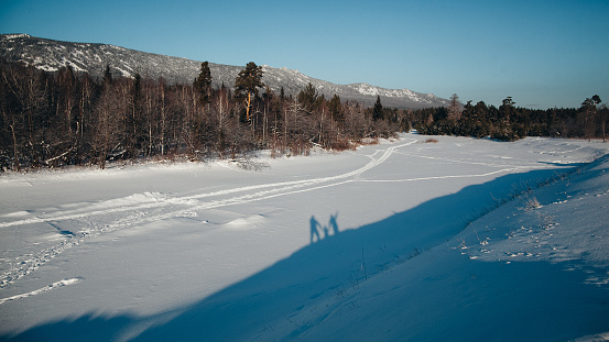 A couple is walking on a frozen lake on a bright winter day. There are snow-covered trees on the shore and snow-capped mountains in the distance. The sky is clear and blue.