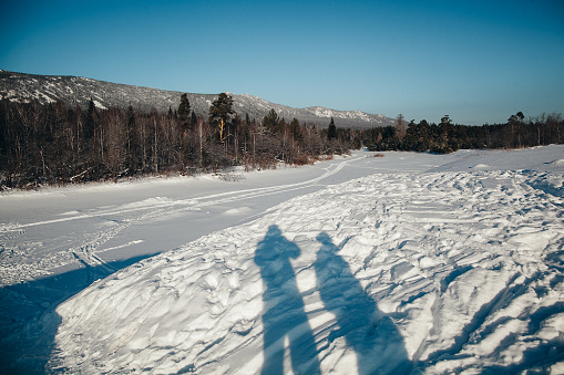 The image shows two people standing on a frozen lake surrounded by snow-covered mountains. There are trees on the shore of the lake, and the sky is clear and blue.