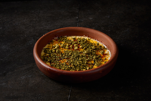 Side view of an elegant crÃ¨me brÃ»lÃ©e with pistachio in a clay bowl, exuding gourmet charm against a stark black backdrop.