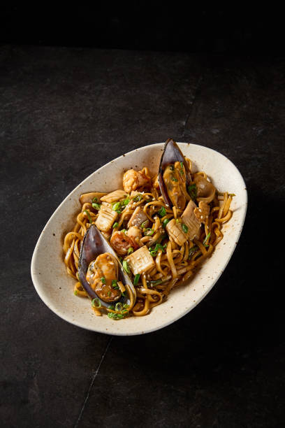 istock Side perspective in a vertical frame showcasing udon pasta, brimming with assorted seafood, served Asian-style on a ceramic plate against a deep black background 1992288664