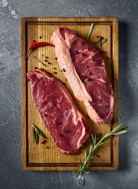 istock Vertical overhead shot, showcasing raw striploin and New York steaks meticulously placed on a wooden board. The marbling of the beef stands out against the gray 1992288696