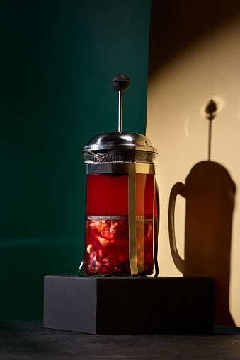 A sophisticated tea creation, encapsulated within a see-through French press, enriched with the vibrant allure of pomegranate seeds, set against a harmonizing black and beige color-blocked backdrop.