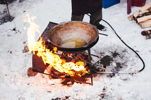 A person in heavy coat and hat stirring a cast iron cauldron filled with meat, vegetables, and spices over a fire on coals with a stack of firewood nearby in a rustic outdoor setting.