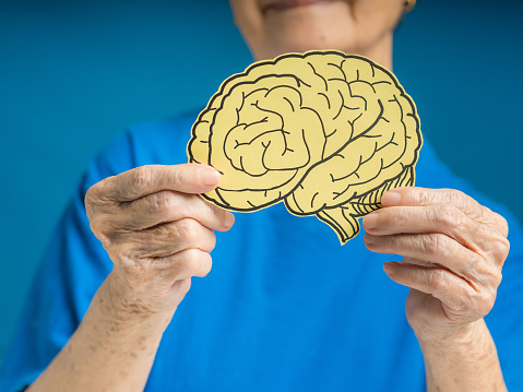Hands of a senior woman holding a brain shape made from yellow paper against a blue background. Alzheimer's, Parkinson's disease, dementia, stroke, seizure, or mental health. Healthcare concept