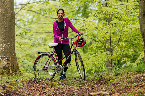 A low-angle full-length shot of a young female adult standing with her bicycle in a wooded area in the countryside of Northumberland, North East England. She is looking off-frame and smiling as she takes a break. She is wearing casual clothing and her bicycle helmet is hanging on the handlebars. Behind her is lush greenery.