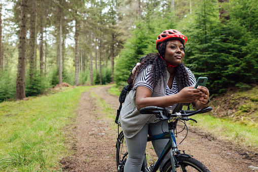 A three-quarter-length shot of a young female adult leaning forwards on her bicycle in a wooded area in the countryside of Northumberland, North East England. She is looking ahead, away from the camera, whilst holding her mobile phone. She is wearing casual clothing and a bicycle helmet, behind her there are large trees and a forest track which curves away.